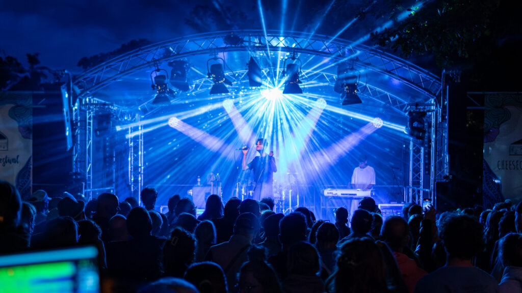 Auf dem Foto ist eine Bühne zu sehen, auf der zwei männliche Musiker stehen. Eine Person singt und die andere Person spielt Keyboard. Vor der Bühne stehen viele Personen mit dem Rücken zum Bild. Die Personen sind das Publikum.