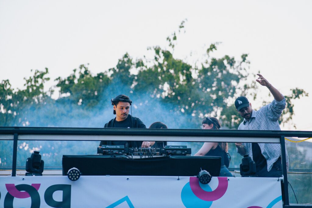 Auf dem Bild sind mehrere Personen zu sehen. Eine Person ist ein DJ. Die Person ist männlich und steht vor einem DJ-Pult. Die anderen Personen tanzen um ihn herum.