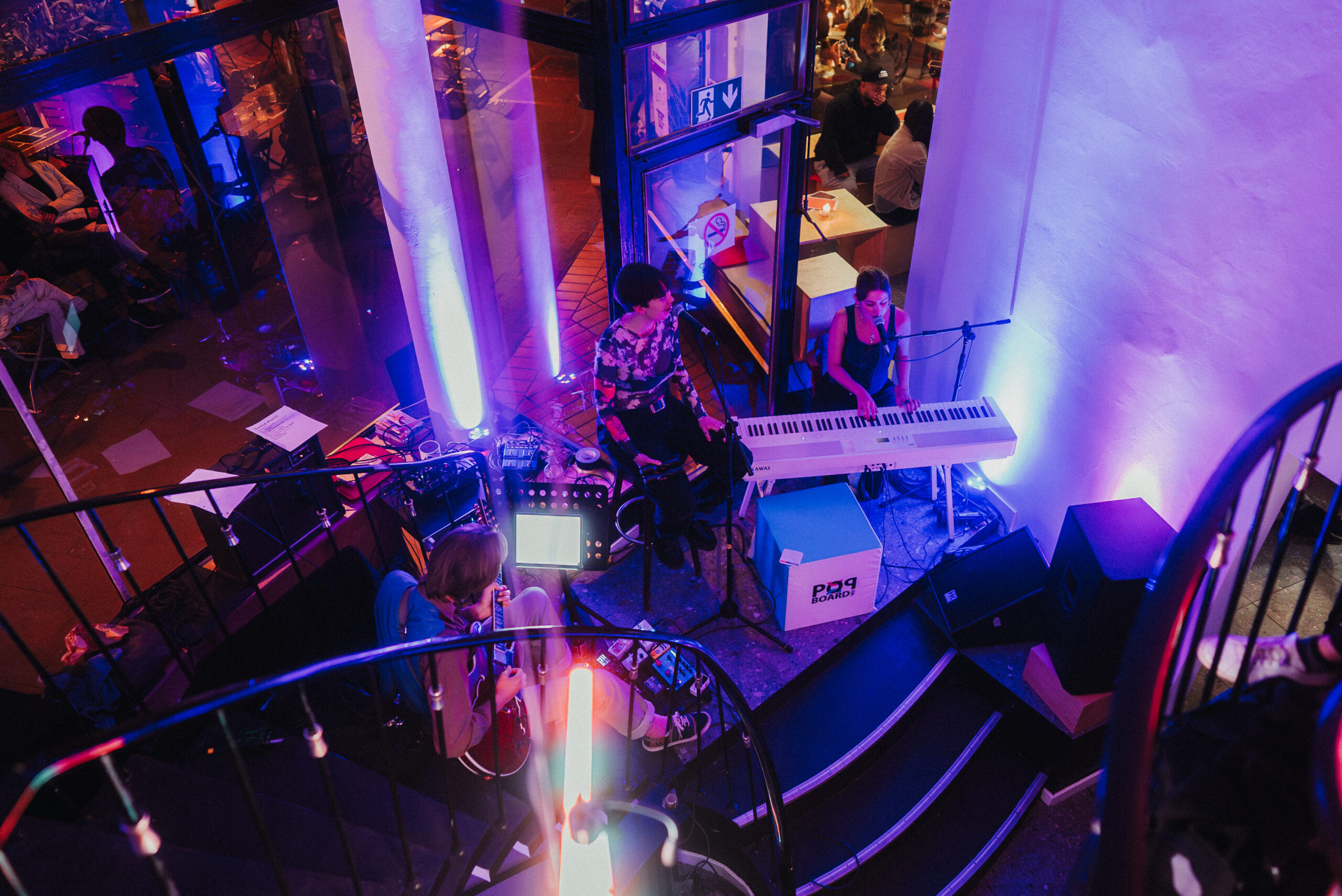 Auf dem Bild sind drei Personen auf einer Bühne zu sehen. Die Personen sind eine Band und spielen verschiedene Musikinstrumente. Das Bild wurde zeigt die Perspektive von oben.