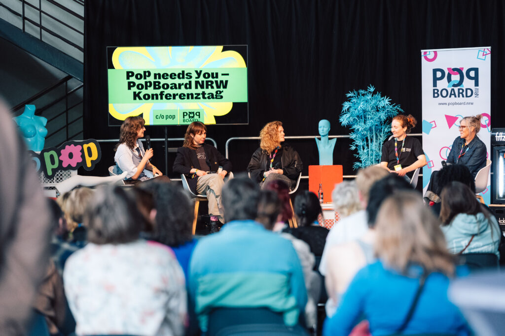 Auf dem Foto sitzen fünf weibliche Personen auf einer Bühne. Eine Person hält ein Mikrofon in der Hand. Im Hintergrund ist ein TV-Screen zu sehen, darauf steht: Pop Needs you – PopBoard NRW Konferenztag". Im Vordergrund sitzt das Publikum vor der Bühne mir dem Rücken zum Bild.