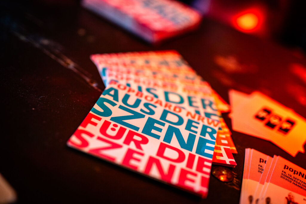Auf dem Foto liegen mehrere Flyer auf einem Tisch. Auf den Flyer steht: Aus der Szene, für die Szene!