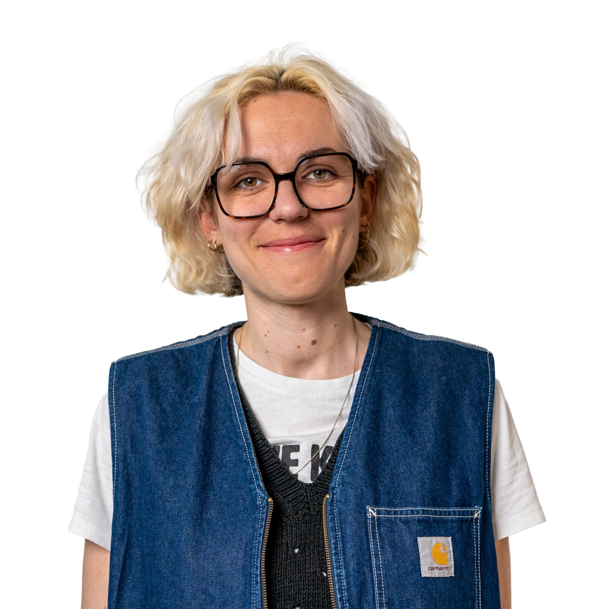 Auf dem Bild ist eine weibliche Person zu sehen die im Porträtmodus in die Kamera blickt. Die Person hat kurze blonde Haare und trägt eine Brille. Die Person hat ein weißes T-Shirt und darüber eine blaue Jeansweste an.