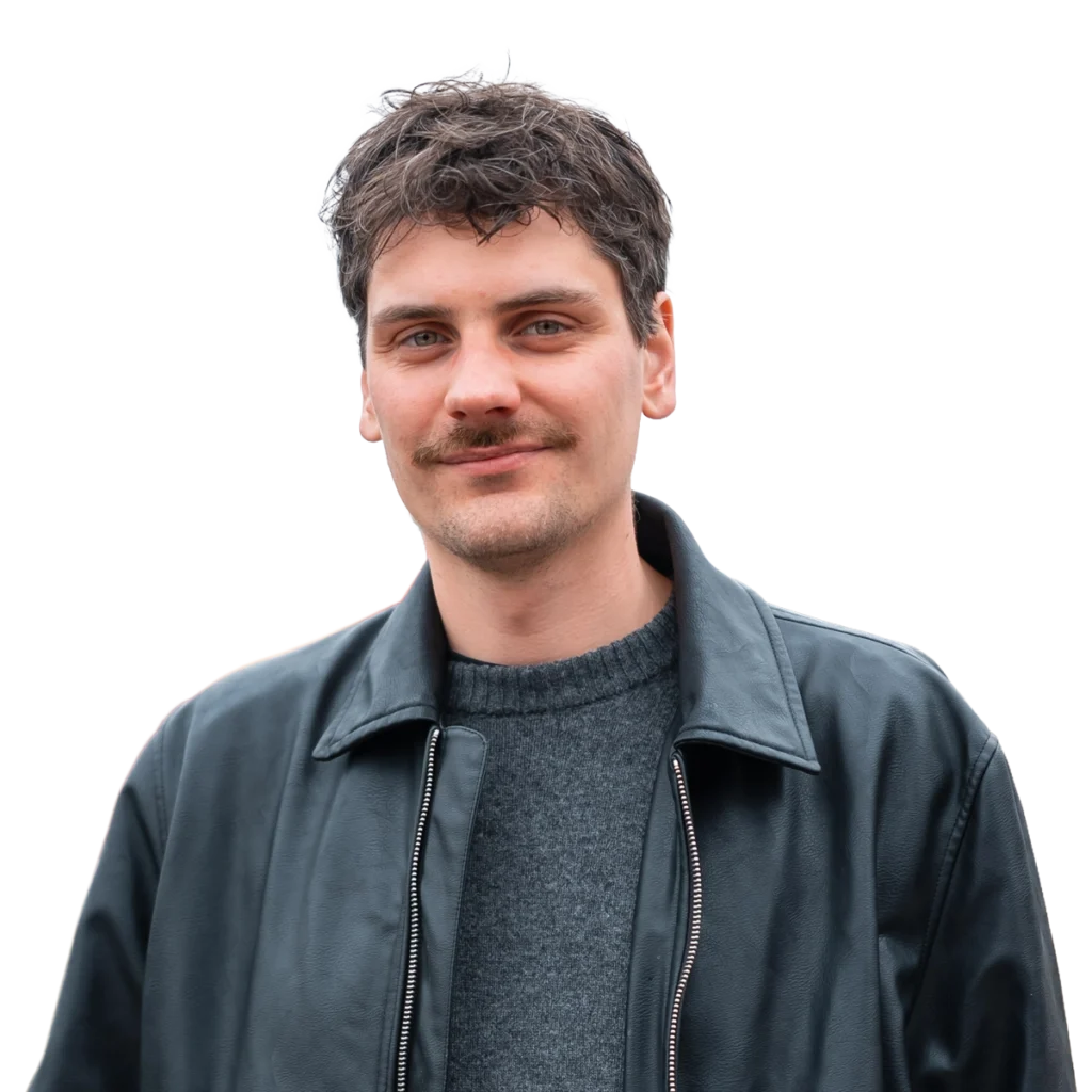 Auf dem Bild ist eine männliche Person zu sehen die im Porträtmodus in die Kamera blickt. Die Person hat kurze braune Haare und trägt eine schwarze Lederjacke. Darunter einen grauen Pullover.