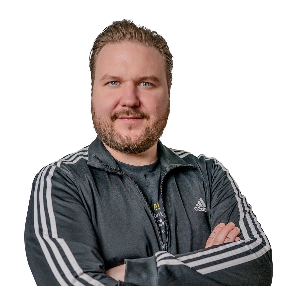 Auf dem Bild ist eine männliche Person zu sehen die im Porträtmodus in die Kamera blickt. Die Person hat hellbraune kurze Haare und einen Bart. Die Person hat einen graue Adidas Trainingsjacke an.